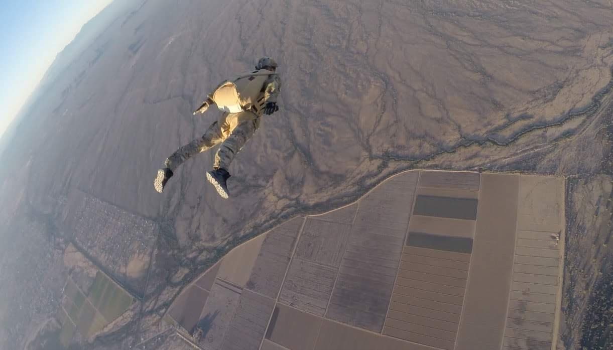 Military HALO free-fall jumper in stable arch position high above desert terrain