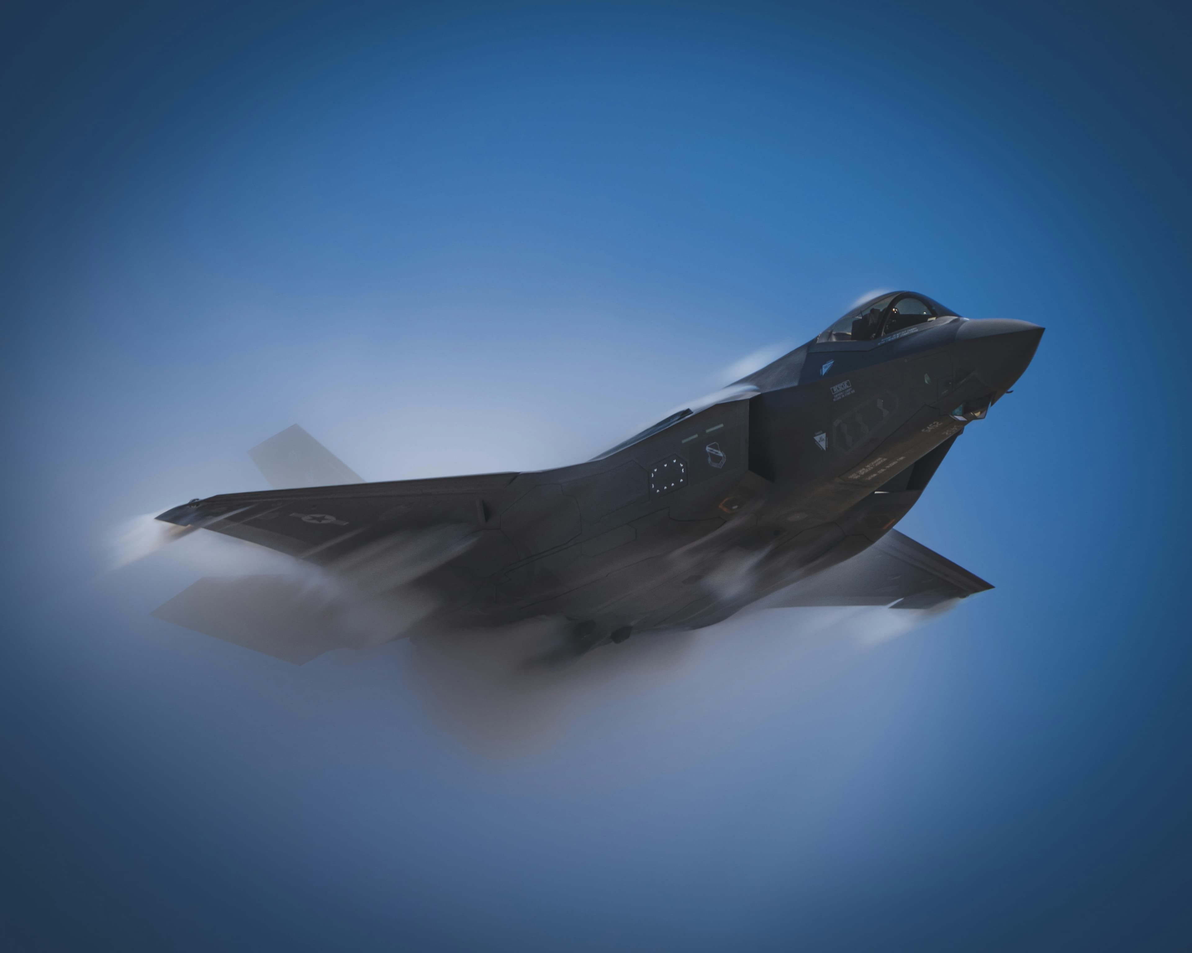 F-35 Lightning II fighter jet banking through a vapor cloud against a deep blue sky